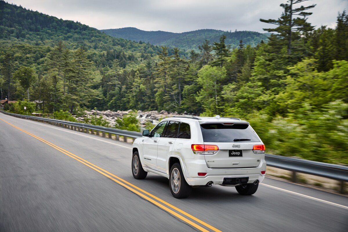 2021-Jeep-GrandCherokee-Overland-SUV-L02.png white Jeep Grand Cherokee SUV driving on a highway through wooded mountains