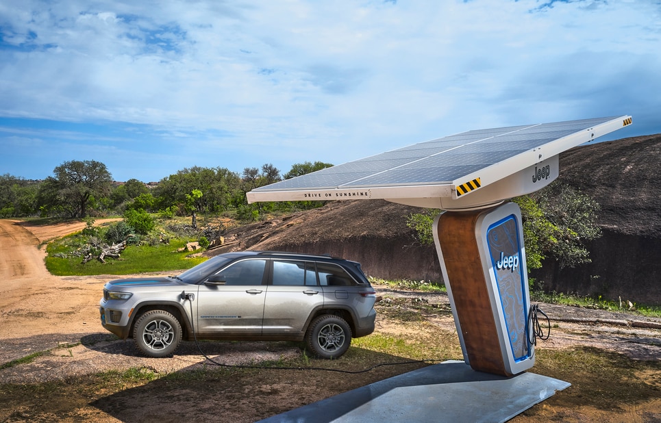 2022-Jeep-GrandCherokeeWL-4xe-Trailhawk-SUV-L12.png silver Jeep Grand Cherokee 4xe hybrid SUV parked under an awning at a charging station