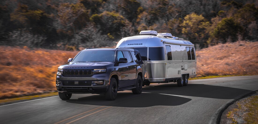 2023-Jeep-WagoneerL-Carbide-SUV-L01.png dark gray Jeep Wagoneer L SUV towing an airstream trailer through a valley