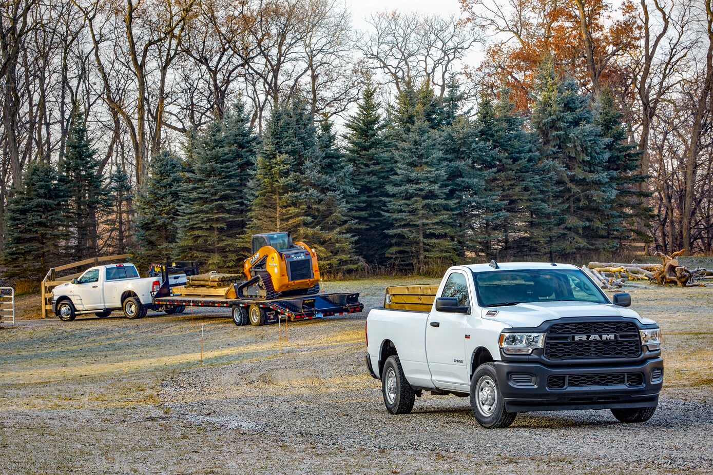 white Ram 2500 truck parked next to another truck, towing a large trailer with a skid loader