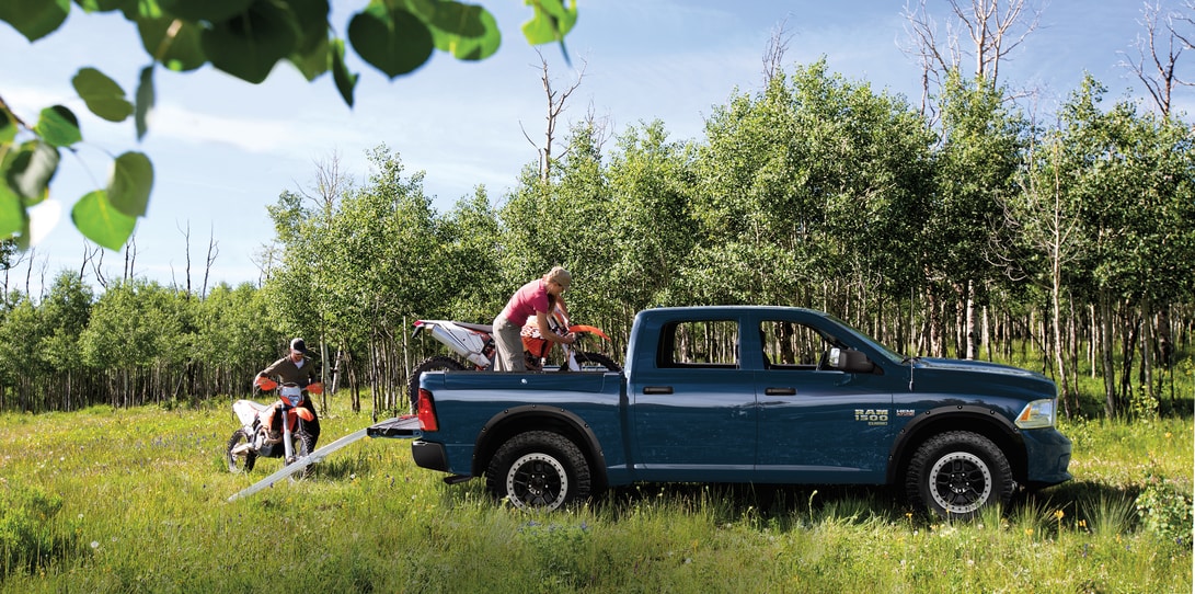 RAM-Lifestyle.png blue Ram 1500 truck parked in a grassy field with a driver and passenger removing motor bikes from the truck bed