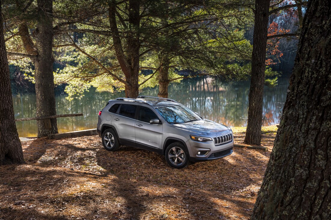silver Jeep Cherokee SUV parked under forest trees next to a stream