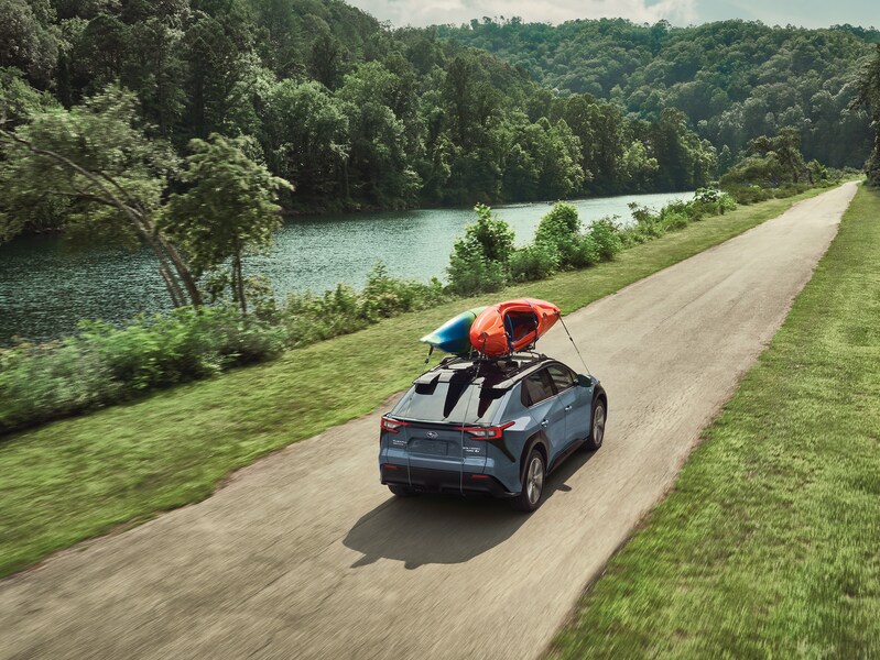 2023-Subaru-Solterra-EV-Touring-SUV.png light blue Subaru Solterra EV driving on a trail near a riverbank with kayaks on the roof rack