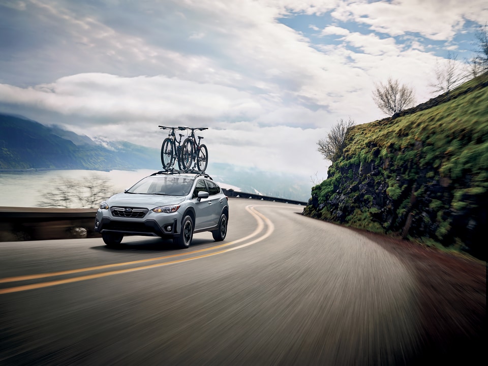2022-Subaru-Crosstrek-Premium-Hatchback-L04.png silver Subaru Crosstrek crossover SUV driving past a waterway with bikes on the roof-rack