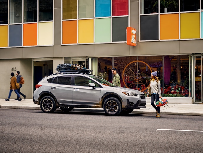 2023-Subaru-Crosstrek-Premium-Hatchback-Crossover.png silver Subaru Crosstrek Crossover SUV parked in front of a camping gear shop with supplies on the roof rack