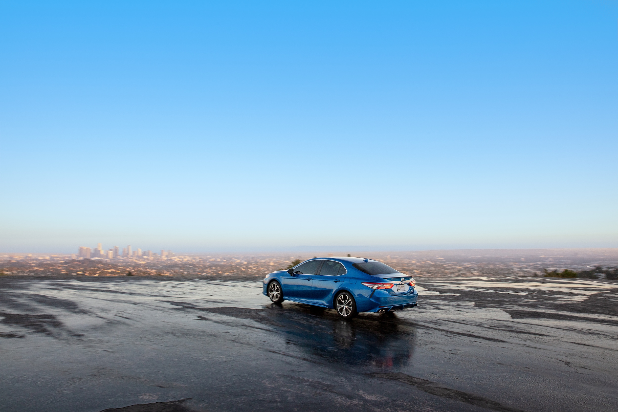 light blue Toyota Camry driving on wet gravel, near a city