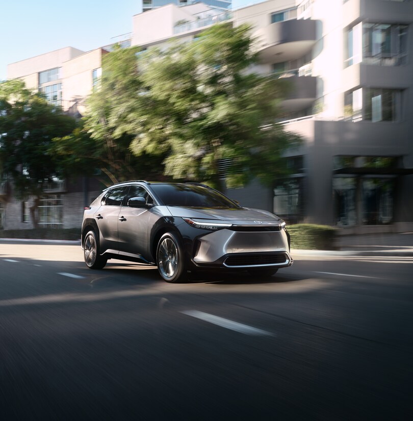 2023-Toyota-bZ4X-Limited-SUV.png silver Toyota bZ4X electric SUV driving down a tree-lined street with an office building in the background