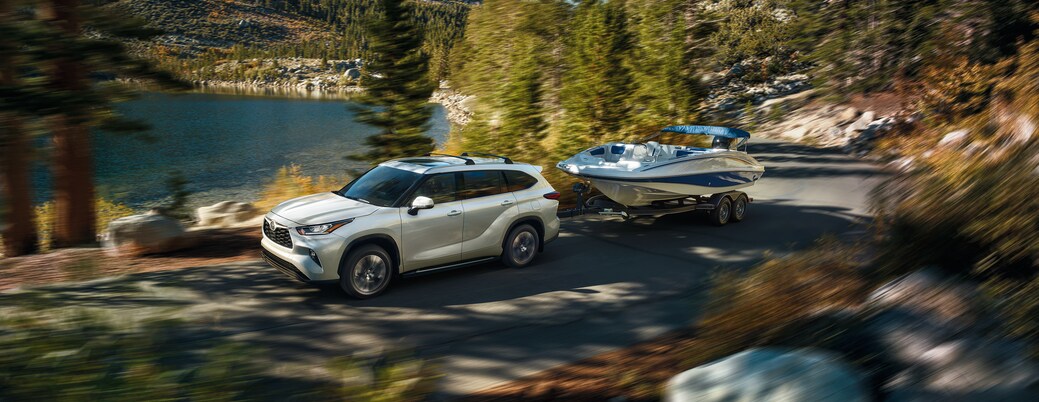 2023-Toyota-Highlander-Platinum-SUV-L05 (2).png white Toyota Highlander SUV driving next to a forest lake, towing a boat on a trailer
