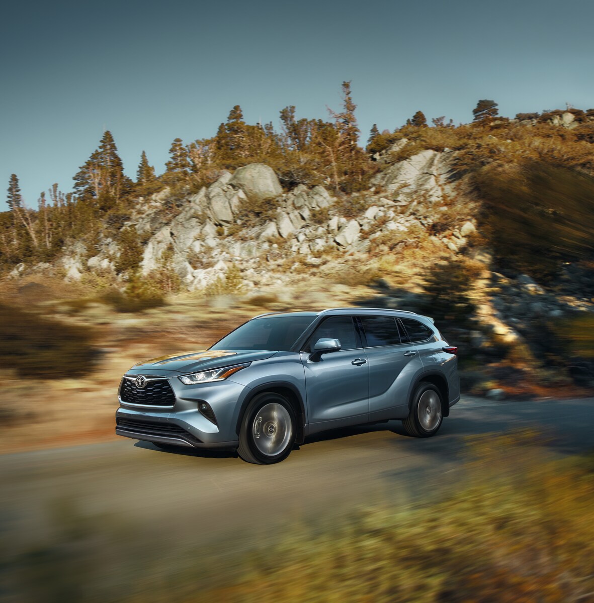 2023-Toyota-Highlander-Platinum-SUV-L06.png silver Toyota Highlander SUV driving down a rocky, tree lined highway
