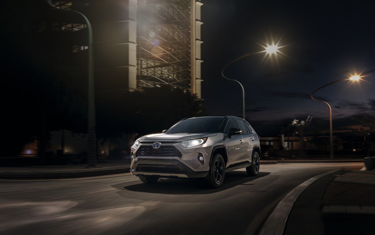 2021-Toyota-RAV4Hybrid-XSE-SUV-L02.png silver Toyota RAV4 Hybrid SUV driving at night through a city with the headlights on