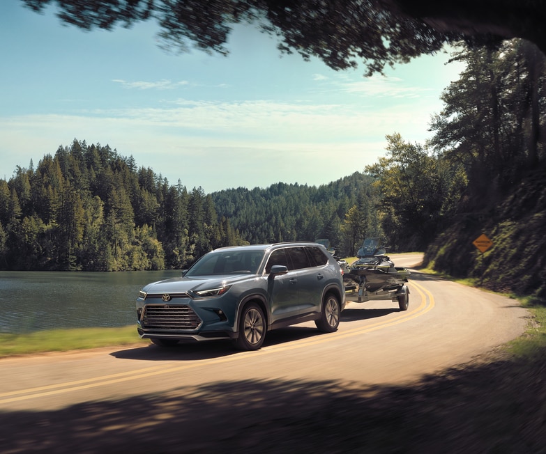 2024-Toyota-GrandHighlanderHybrid-PlatinumMax-SUV.png blue grey Toyota Grand Highlander Hybrid driving next to a lake, towing a skidoo