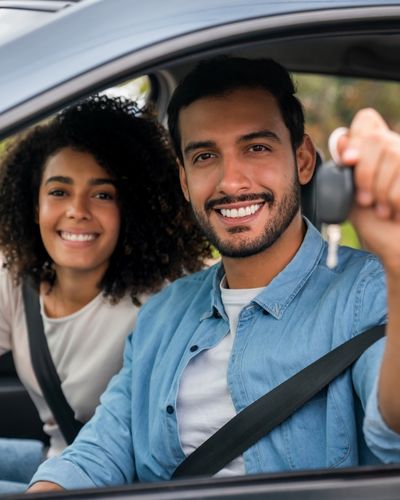 New%20Car%20Owners.jpg Hispanic man holding up key in new car with African-American woman sitting in the passenger seat smiling