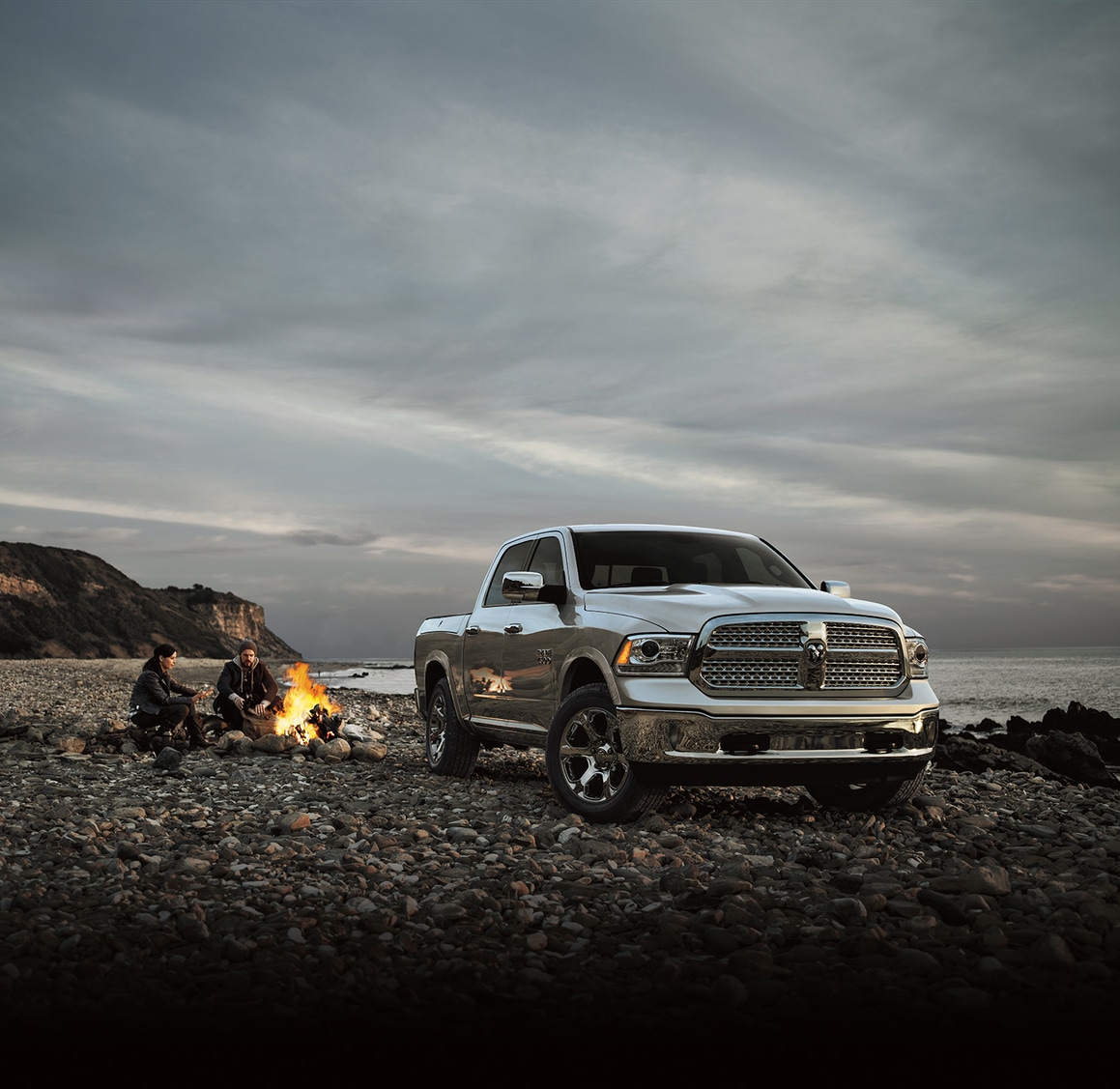 2015-Ram-1500-Laramie-CrewCab.png silver Ram 1500 truck parked on a rocky beach with a couple and campfire nearby