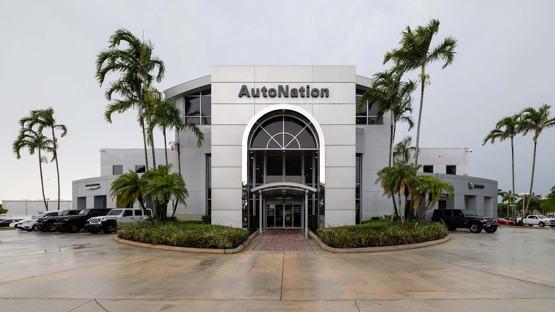 Exterior view of AutoNation Chrysler Dodge Jeep RAM Pembroke Pines during the day. There is a cloudy sky and many vehicles parked near the grey building. Trees and greenery are visible around the parking lot.