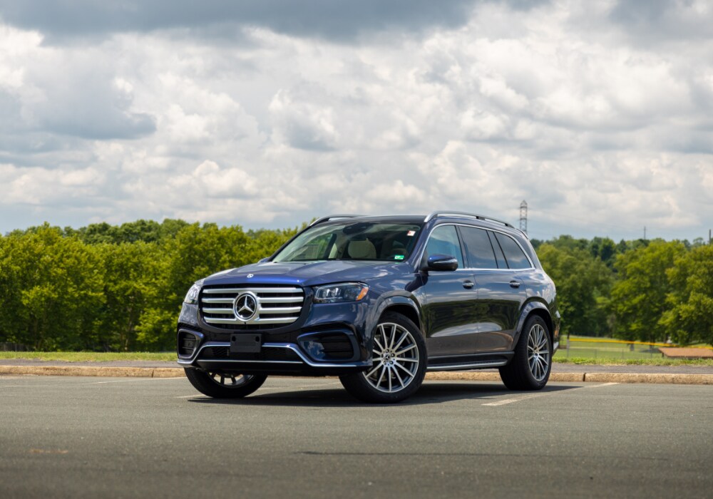 mercedes-gls-450-for-sale.png Dark blue 2025 Mercedes-Benz GLS 450 SUV for sale parked in a paved lot with trees and clouds in the background