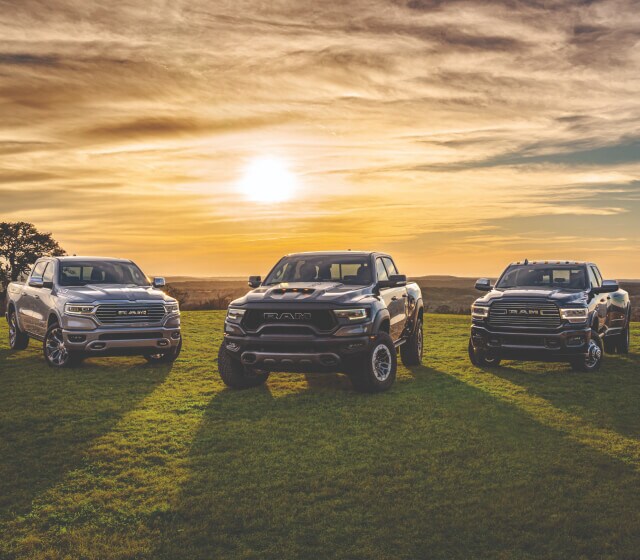 Ram 1500 Limited Longhorn in Billet Silver, Ram 1500 TRX in Granite Crystal, and a Ram 3500 Laramie in Maximum Steel parked on a grassy hill with the sun setting in the background