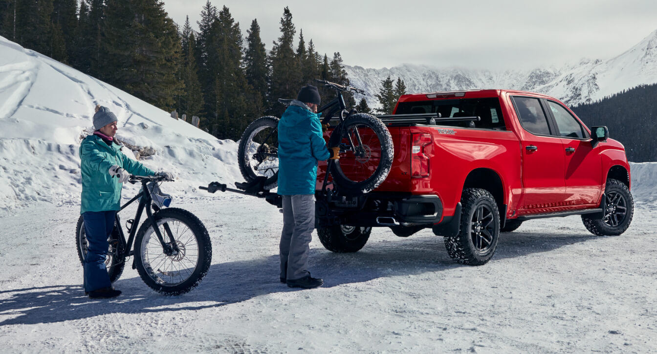 Two people in snow gear unload mountain bikes from a Chevy Silverado bike rack on a snowy mountain road.