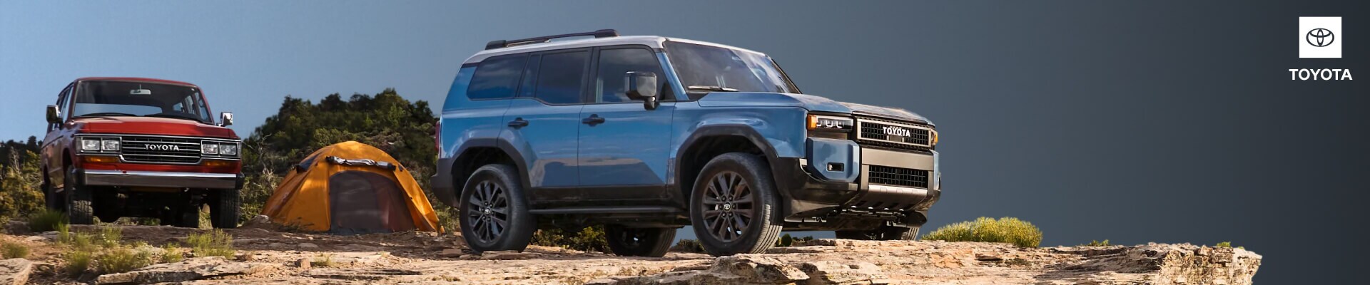 Front three-quarter view of a blue Toyota Land Cruiser alongside a vintage red Land Cruiser on a rocky cliffside campsite.