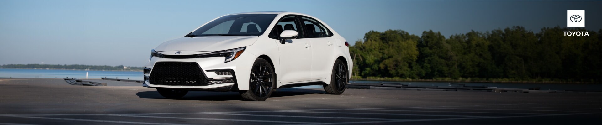 Front three-quarter view of a 2027 Toyota Corolla XSE in White Chill Pearl with gloss-black alloy wheels, parked near a waterfront dock.