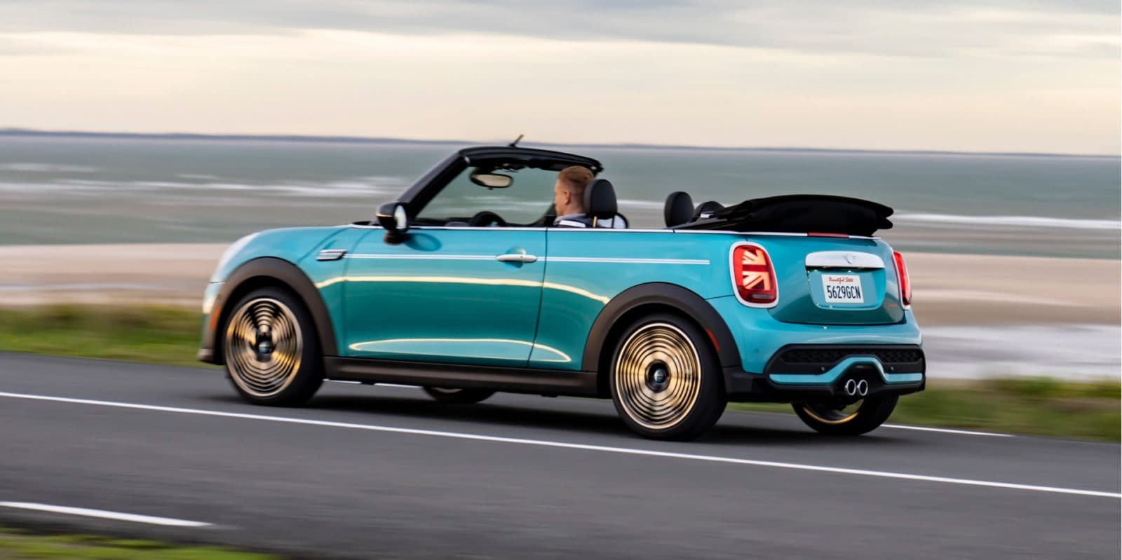 2.jpeg Side view of a MINI Cooper S Convertible Seaside Edition in Caribbean Aqua driving along a coastal road with a man in the driver's seat and no one else in the vehicle, including a beach and cloudy skies in the background.