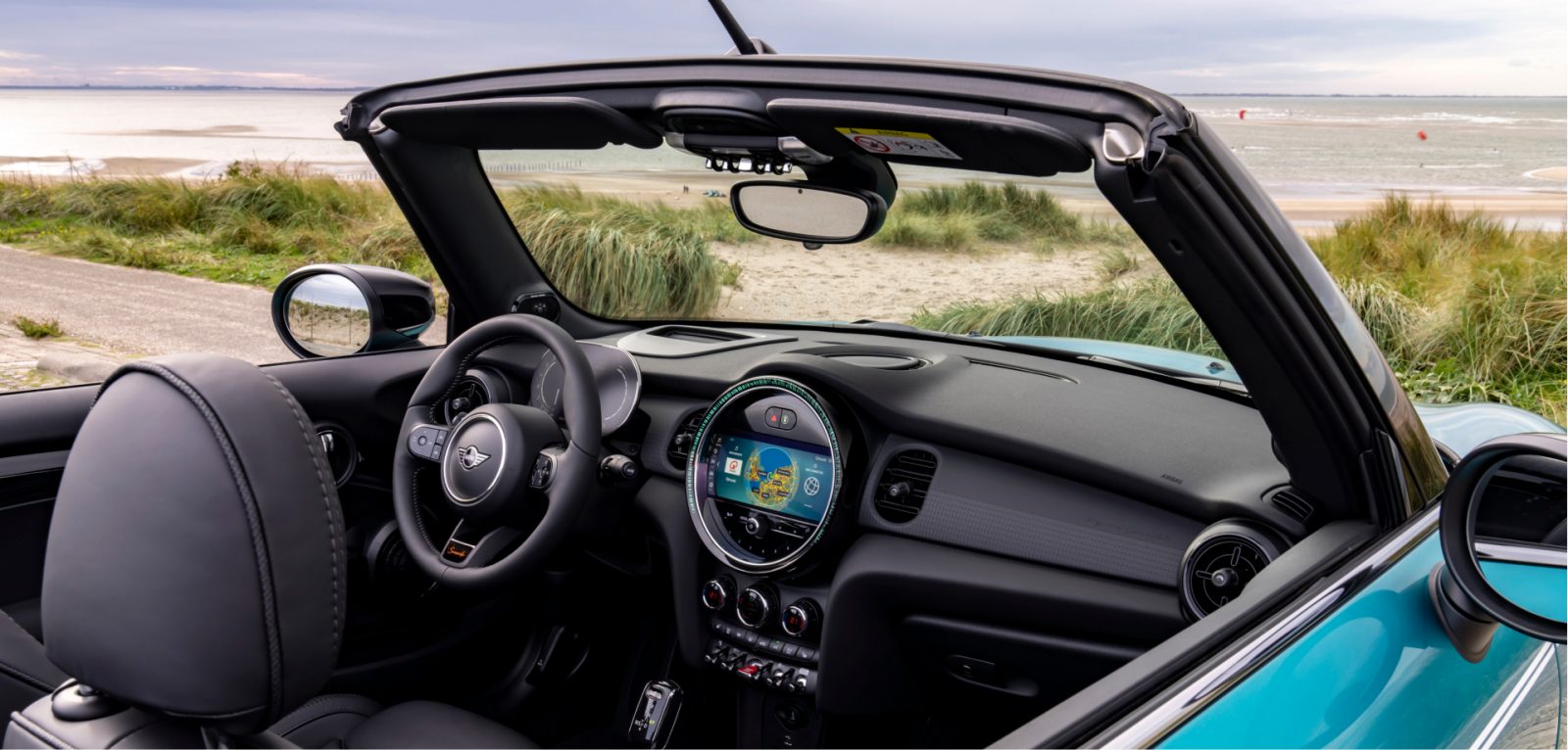 7.jpeg Closeup view of the front row of a MINI Cooper S Convertible Seaside Edition in Caribbean Aqua, with the vehicle's top down and a coastal view in the background including dune grass plus cloudy skies