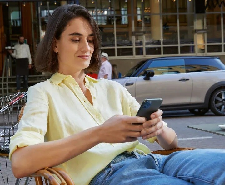 2. Alt. Female wearing yellow collared button-down shirt and jeans, sitting in a chair and holding a smartphone.jpg Woman wearing yellow collared button down shirt and jeans, sitting in a chair and holding a smartphone next to a street with a grey MINI vehicle parked in the background.