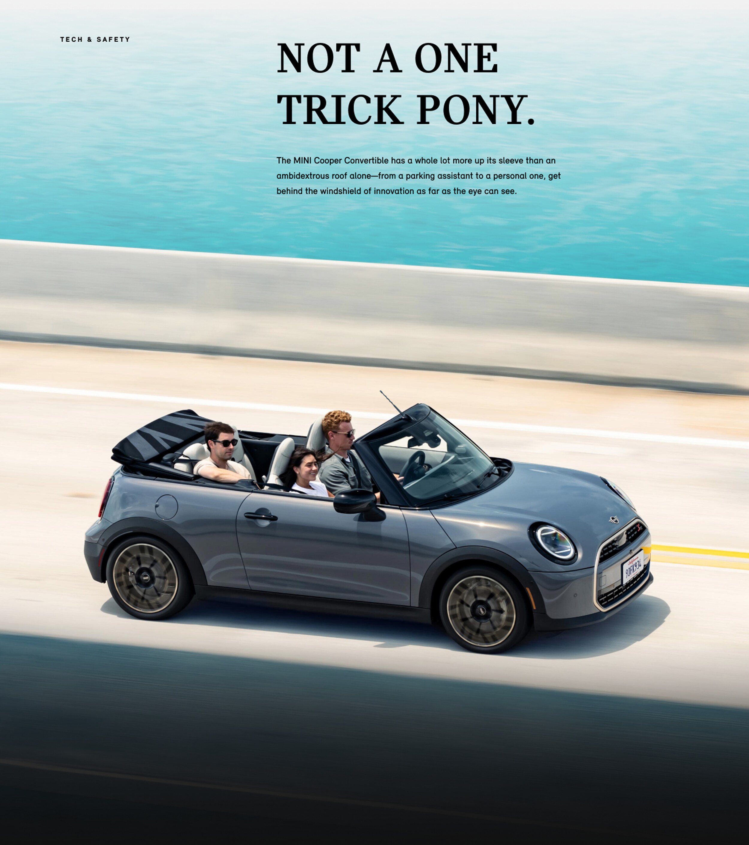 Grey MINI Cooper Convertible with three passengers driving on a seaside highway.