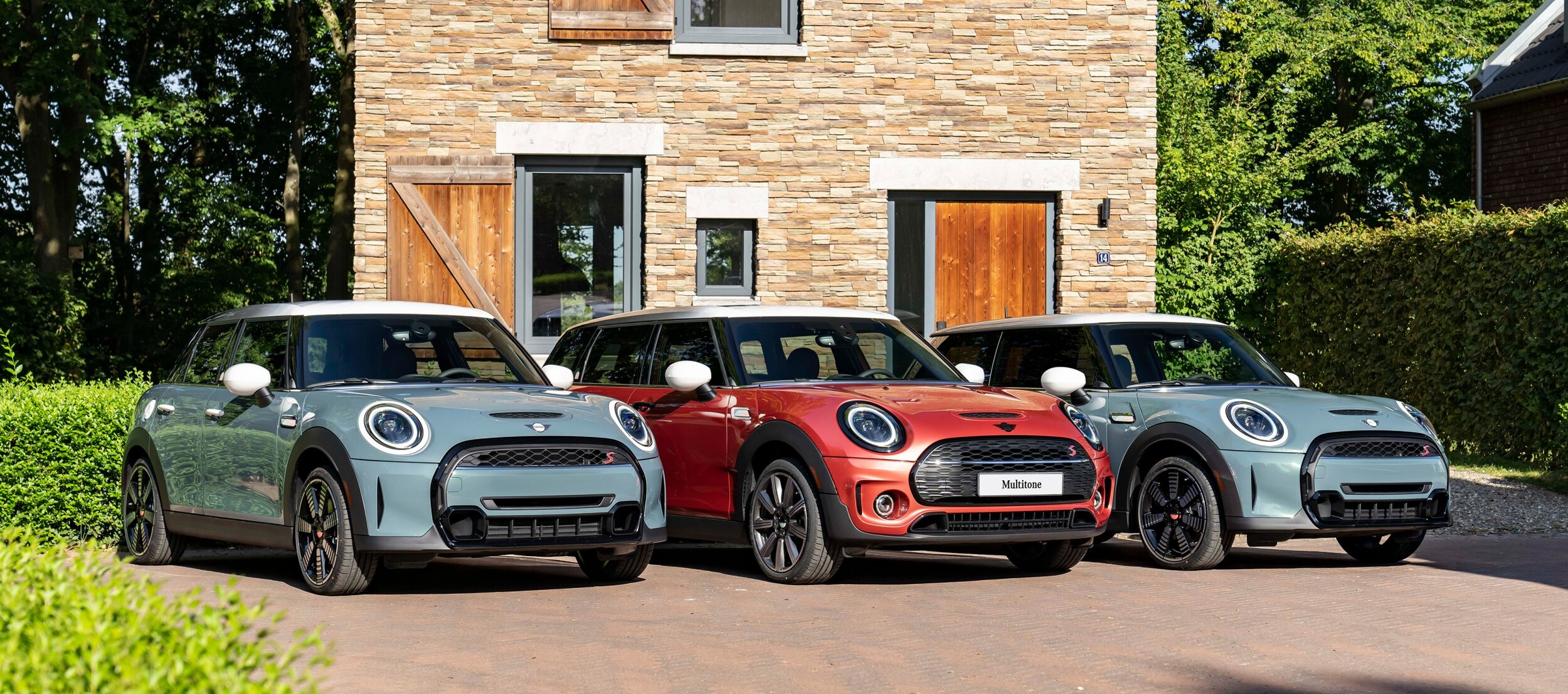 1.jpg Three MINI Multitone Edition vehicles, two in Sage Green and one in Coral Red, parked side by side on a brick surface in front of a brown brick building with greenery surrounding it.