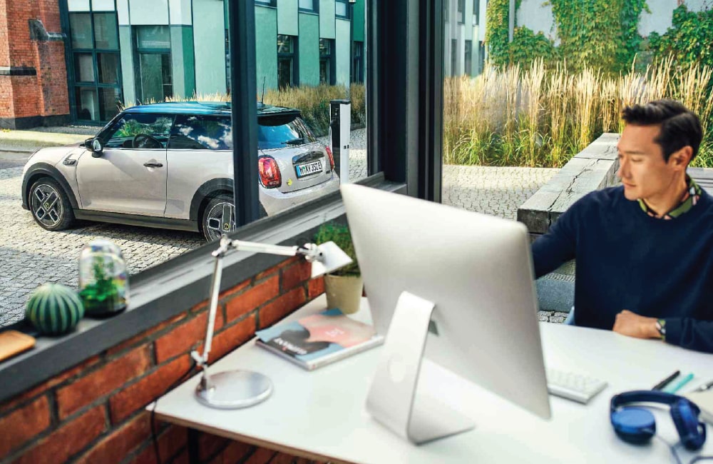 Image-10.png Man sitting at desk on the computer with a MINI parked on the street outside behind him