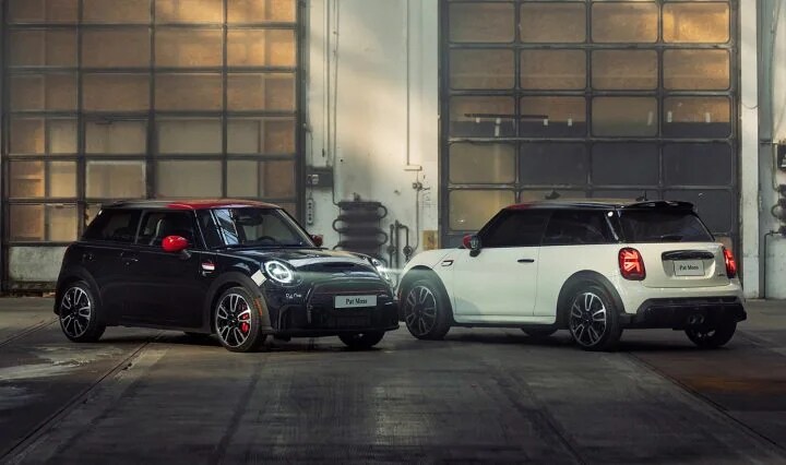 Two MINI Pat Moss Edition vehicles side by side, the left one (Midnight Black metallic) in a three-quarter front view and the right one (Pepper White) in a three-quarter back view, parked on a cement surface in a warehouse type space.