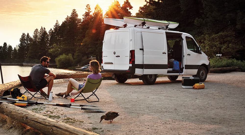 Two people sitting by a lake near a rear angle view of a white 2025 Mercedes-Benz Sprinter 2500 Cargo Van.