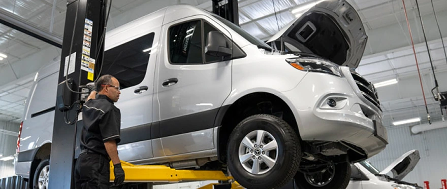 Technician working beneath a white Sprinter van raised on a workshop lift