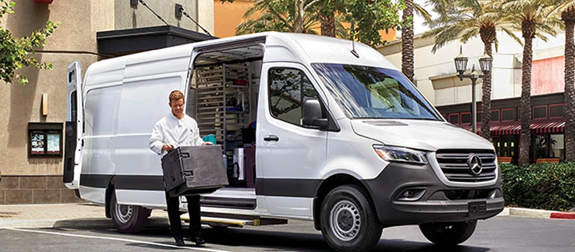 Chef lifting a crate from the open side door of a white Sprinter Cargo Van parked outside a restaurant