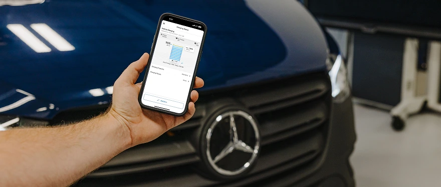 Hand holding a phone showing a charge-level chart in front of the grille of a dark Mercedes-Benz van