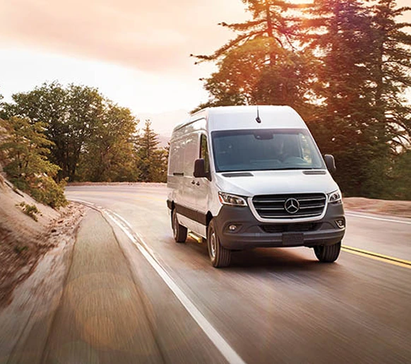 White Sprinter Cargo Van driving down a tree-lined country road into low evening sun
