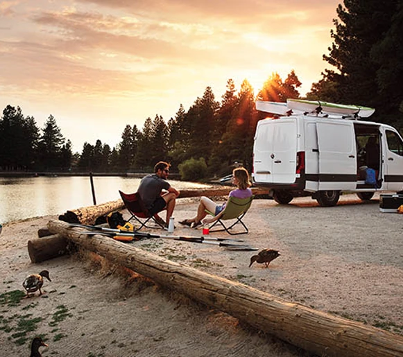 Two people in camp chairs beside a Sprinter van on a lakeshore at sunset