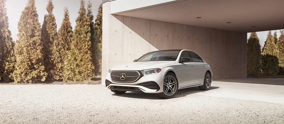 White E-Class Sedan parked beside a modern house with tall trees behind