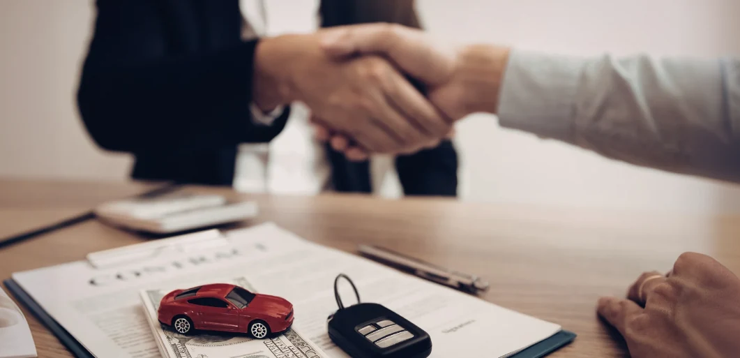 Two people shaking hands over a desk holding a signed contract, car keys and a red toy car