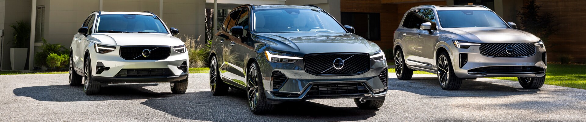 Front and three-quarter view of Volvo XC40, XC60, and XC90 SUVs parked on a driveway outside a modern luxury residence.