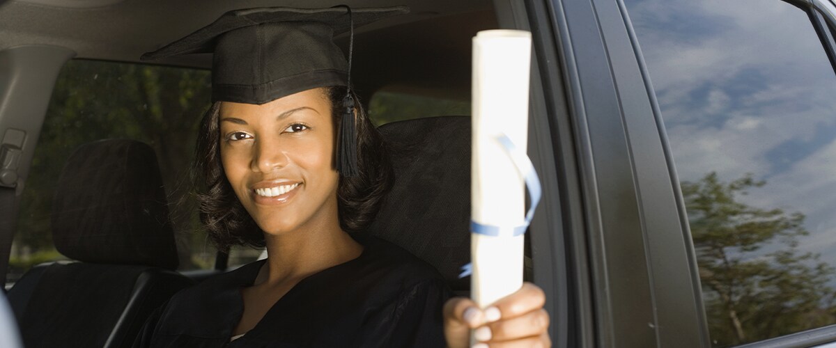 College Grad in cap and gown holds out diploma while sitting in a car