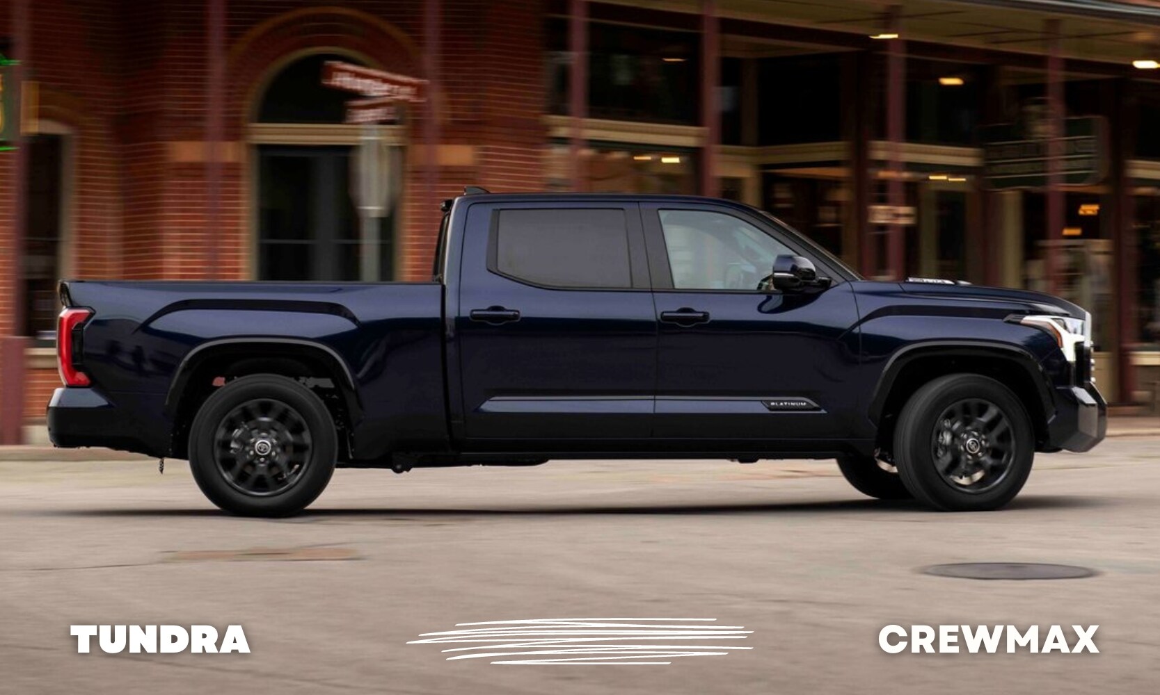 Passenger side view of a blue used Toyota Tundra truck with a CrewMax cab configuration driving through a downtown city street