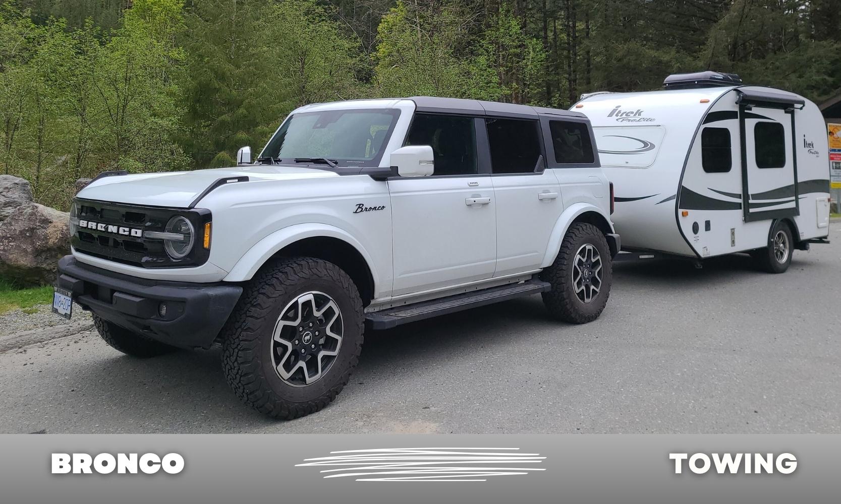 Driver side angle of a white Ford Bronco with a black hardtop roof towing a black and white trailer