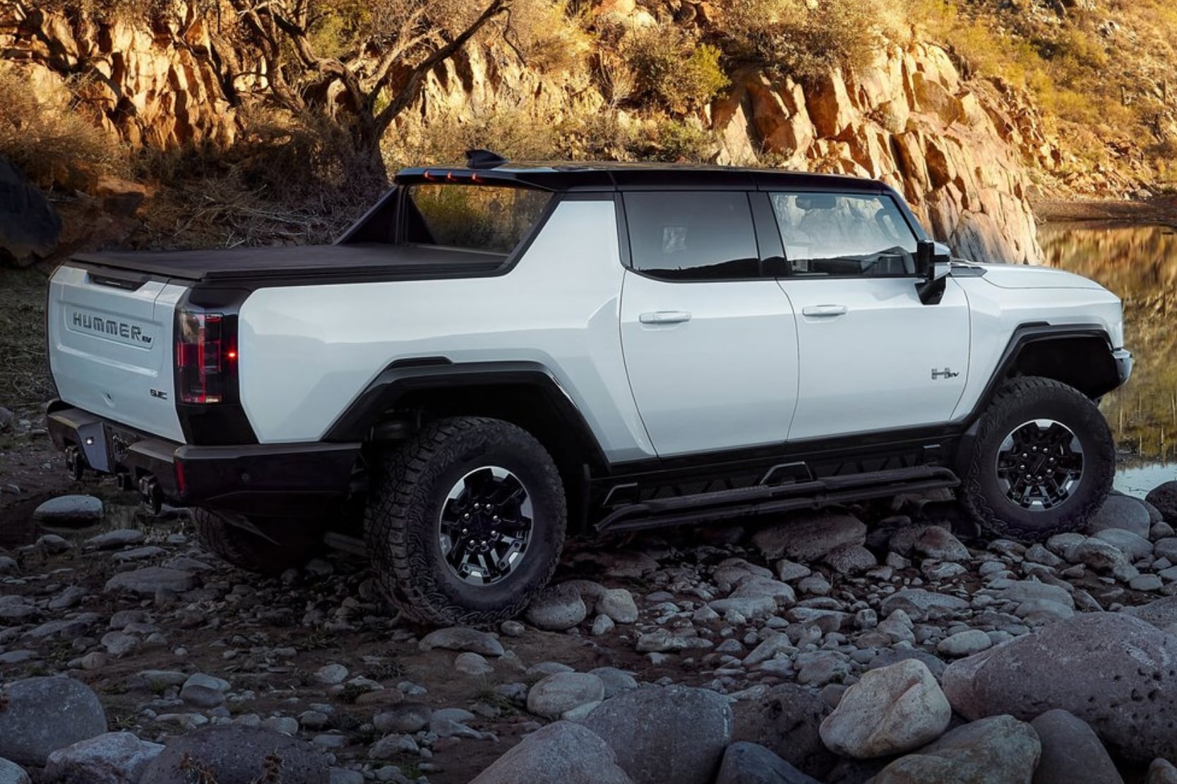 Rear exterior profile view of a white 2024 GMC Hummer EV Pickup Truck parked on some rocks by a lake