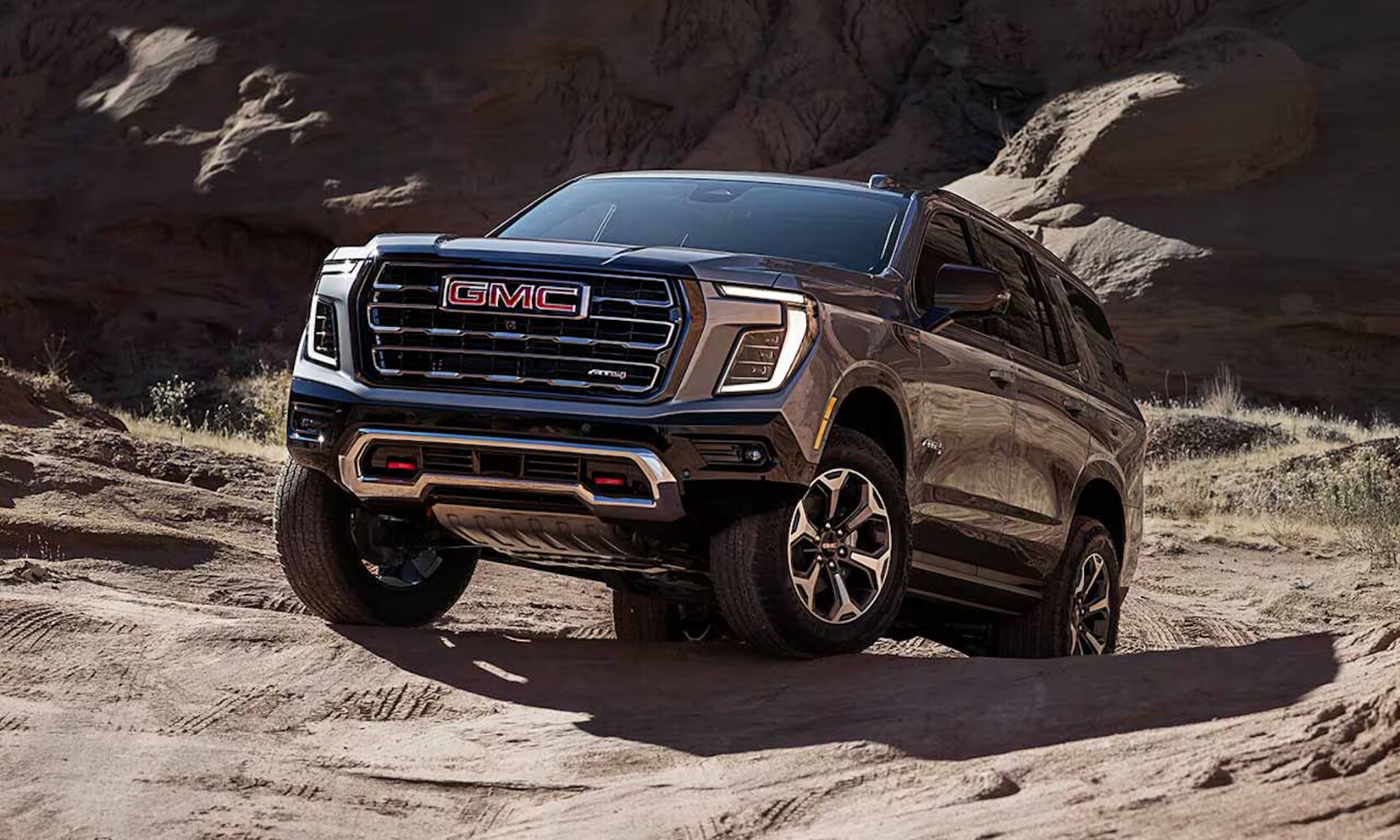 2026%20GMC%20Yukon%20Engines%20-%20Octane%20GMC%20of%20Santa%20Fe.png Exterior front image of a 2026 GMC Yukon AT4 off-roading up a dirt hill with rocky cliffs behind it