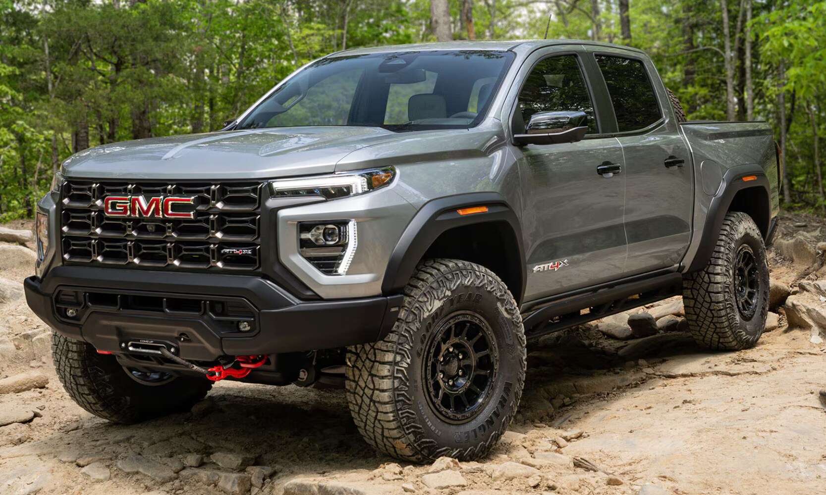 Exterior front angle image of a gray 2026 GMC Canyon AT4X AEV Edition parked in a rocky forest landscape