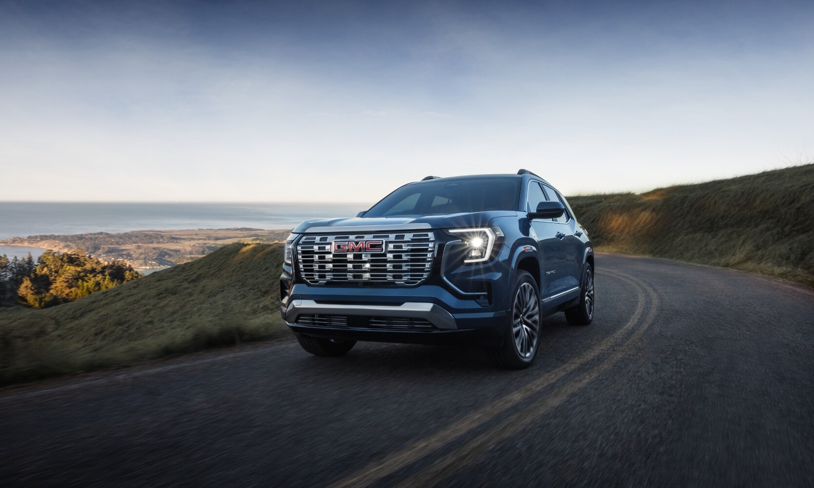 2026%20GMC%20Terrain%20Denali%20-%20Octane%20GMC%20of%20Santa%20Fe.jpg Exterior front image of a blue 2026 GMC Terrain Denali driving along a coastal highway during the day