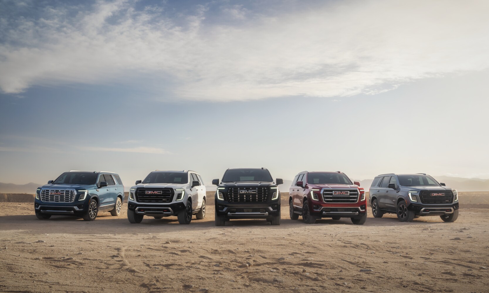 Exterior front image of all five 2026 GMC Yukon trim levels parked in an open dirt field