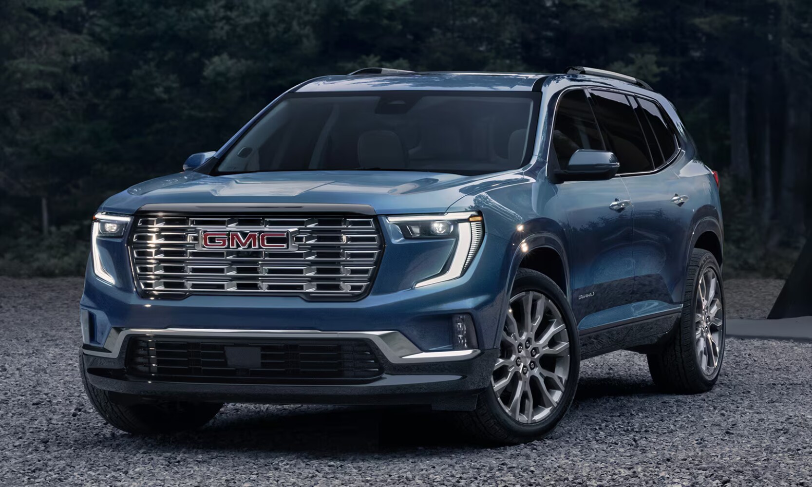 2026%20GMC%20Acadia%20FAQs%20%20Octane%20GMC%20of%20Santa%20Fe.png Exterior front angle image of a blue 2026 GMC Acadia Denali Ultimate parked on a rocky road with forest background views
