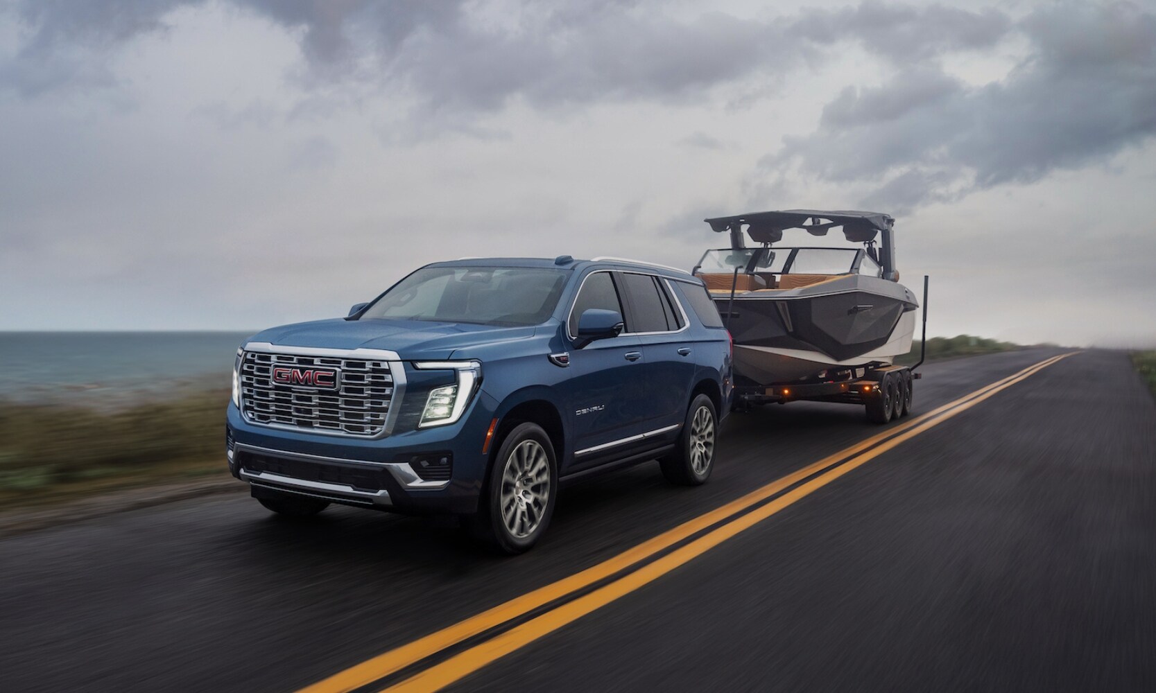2025%20GMC%20Yukon%20vs.%202025%20Chevrolet%20Tahoe%20-%202025%20GMC%20Yukon%20Towing%20Comparison%20-%20Octane%20GMC%20of%20Santa%20Fe.jpg Exterior image of a blue 2025 GMC Yukon towing a small boat on a freeway with water in the background on a cloudy day