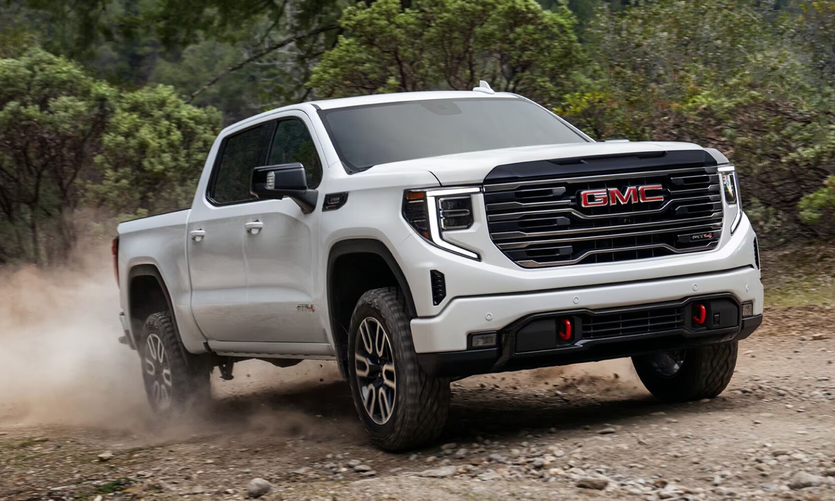 2025%20GMC%20Sierra%201500%20vs.%202025%20RAM%201500%20-%202025%20Sierra%201500%20AT4%20-%20Octane%20GMC%20of%20Santa%20Fe.jpg Exterior image of a white 2025 GMC Sierra 1500 AT4 driving up a rocky dirt road in a forest setting